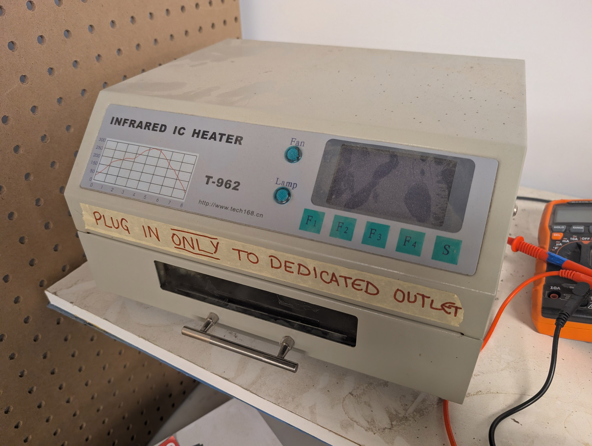 A beige metal box with an angled front top area. The left side of the angled section says "Infrared IC Heater T-962" with a little graph of a reflow temperature printed on, to the right are aqua coloured lamps for Fan and Lamp, and a display to the right of that with F1-F4 and S buttons below it. Masking tape below the angled section says "Plug in ONLY to dedicated outlet" and there is a handle below that to pull a drawer out